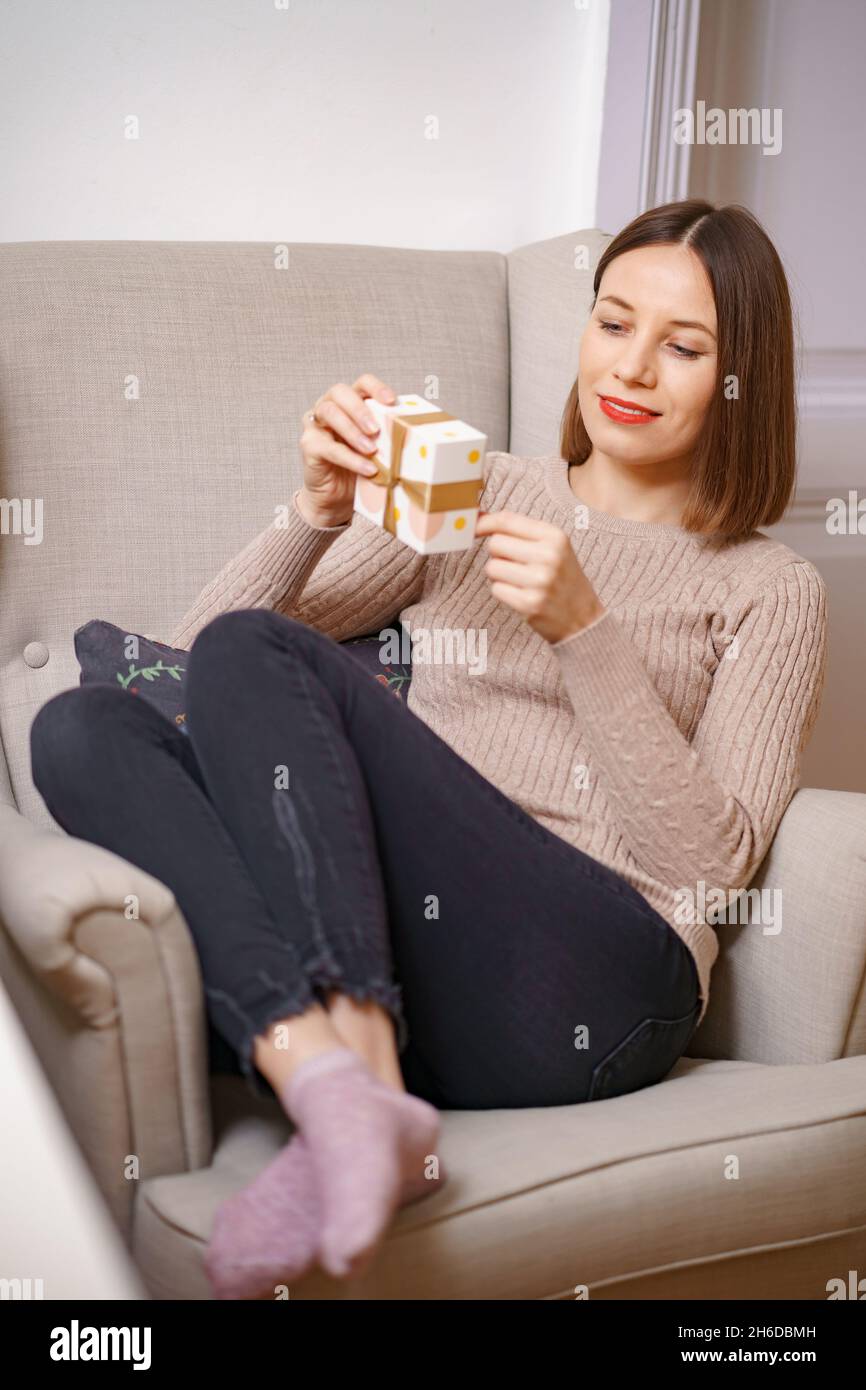 PRETTY young woman while sitting in a comfortable armchair holding a gift box at home. Ghe guesses what's inside Stock Photo