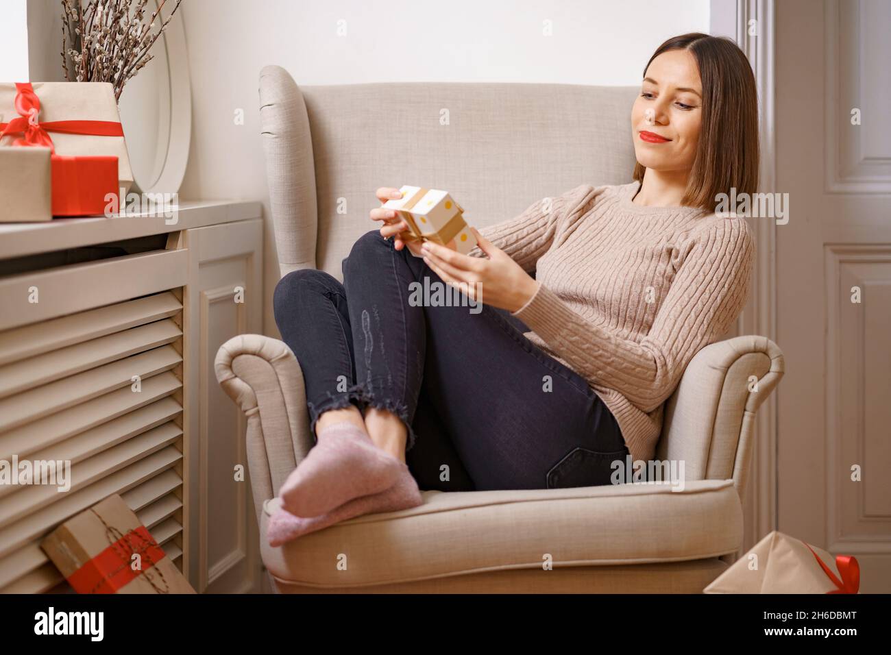 PRETTY young woman while sitting in a comfortable armchair holding a gift box surrounded by gifts at home. Ghe guesses what's inside Stock Photo