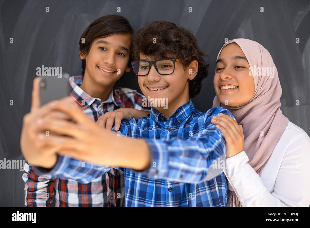 Group of arab teens taking selfie photo on smart phone with black chalkboard in background. Selective focus  Stock Photo