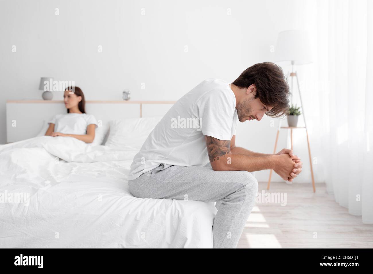 Upset offended caucasian young husband sitting in bed, suffering from weakness, wife ignoring boyfriend in white bedroom Stock Photo