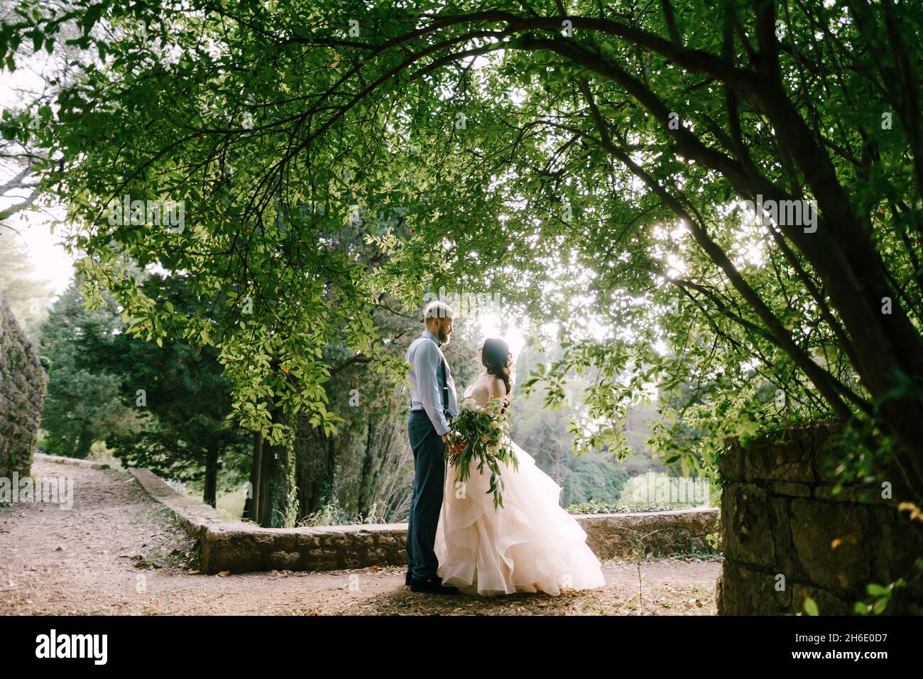 The bride and groom stand holding hands under the lush bush in the ...