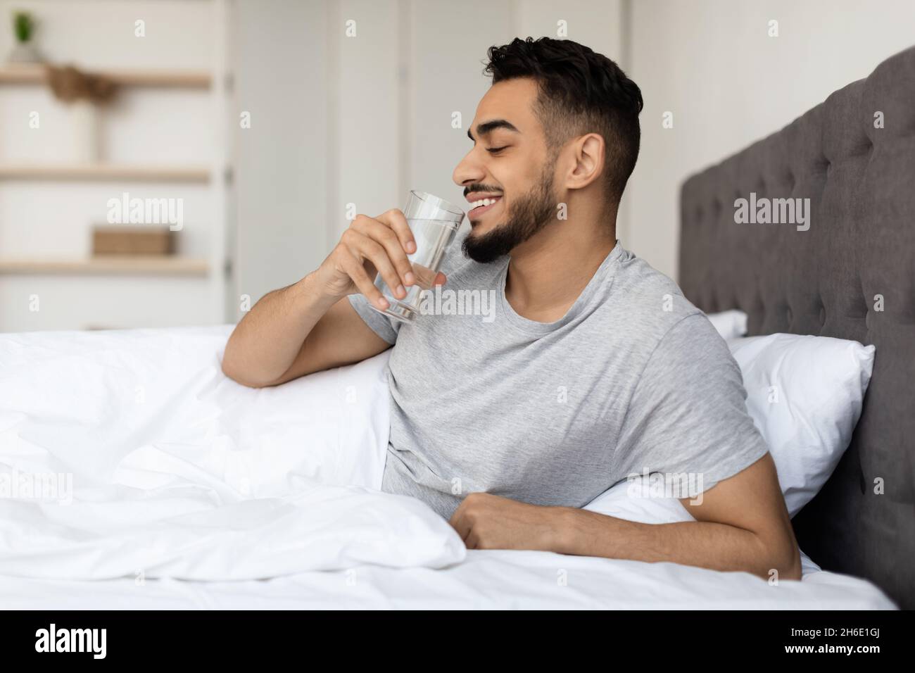 Smiling Arab Guy Drinking Mineral Water From Glass While Sitting In Bed Stock Photo