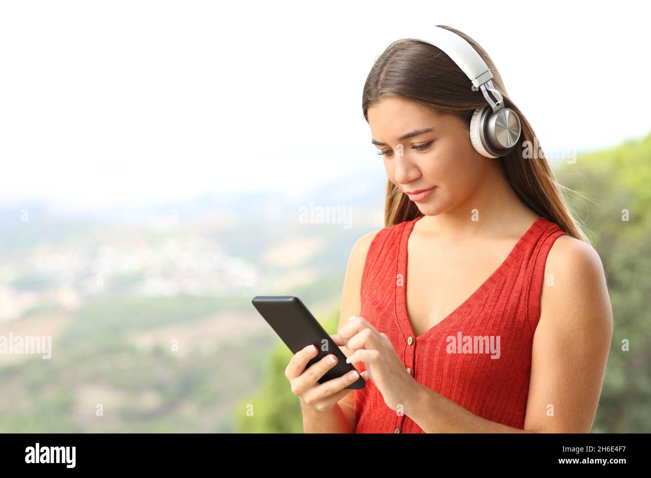 Teenager in red wearing headphones listening to music checking phone outdoors Stock Photo