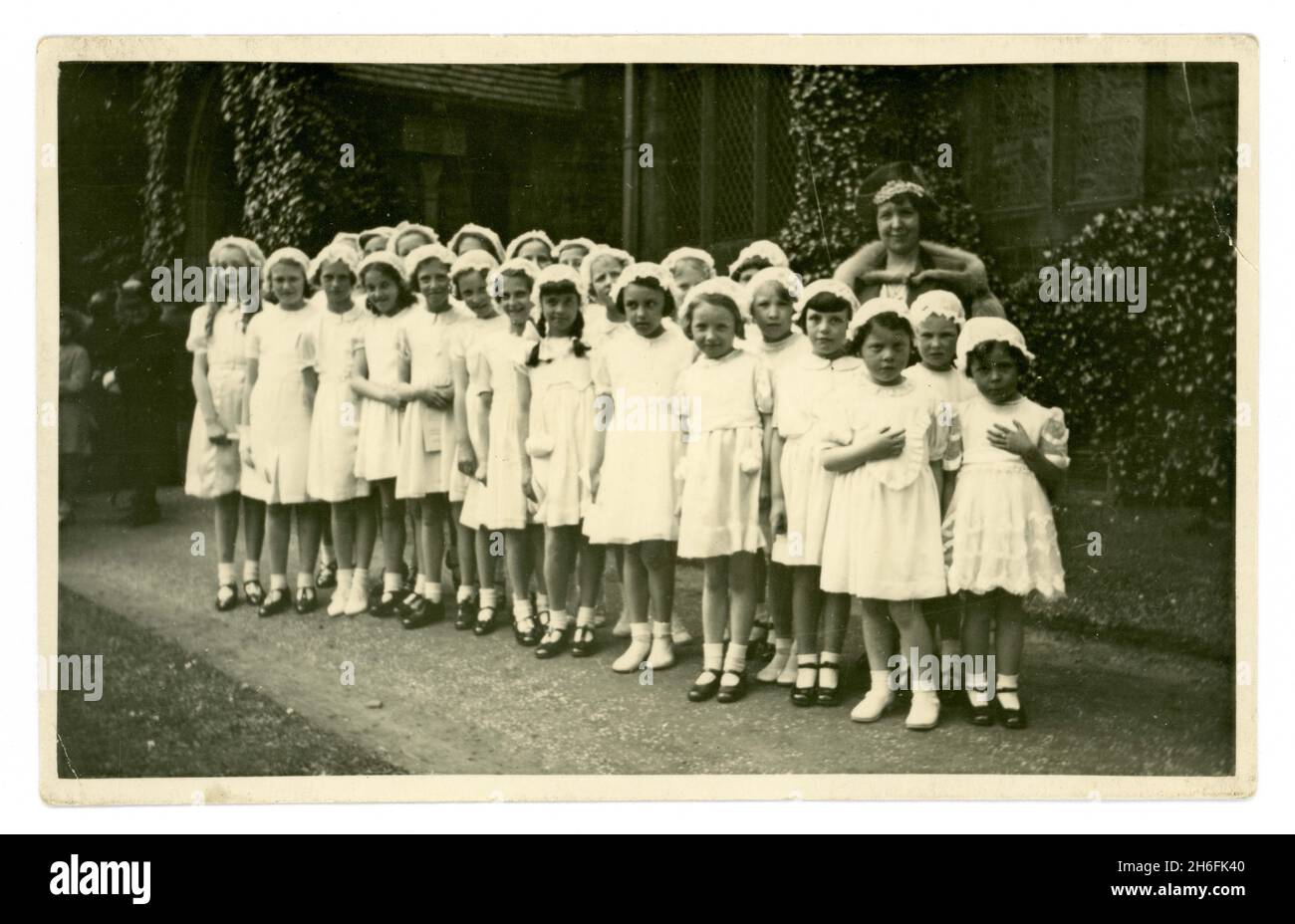 Original 1940's era postcard of young girl's Walking Day. The children are standing in the grounds of a church, they are possibly Sunday School attendees. The young girls are all dressed in white with lace caps, teacher in background in fur stole written on reverse is Miss E. Riley, 37 Rydal Road, church is Christ Church, Heaton, Rydal Road, Bolton, Lancashire, U.K. Stock Photo