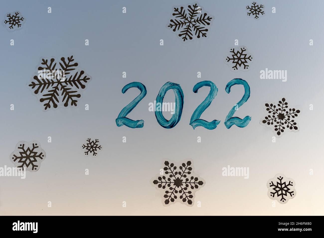 Christmas home window decoration with white snowflakes stickers and handwritten inscription 2022 in blue paint against the setting sun. Snowflake patt Stock Photo