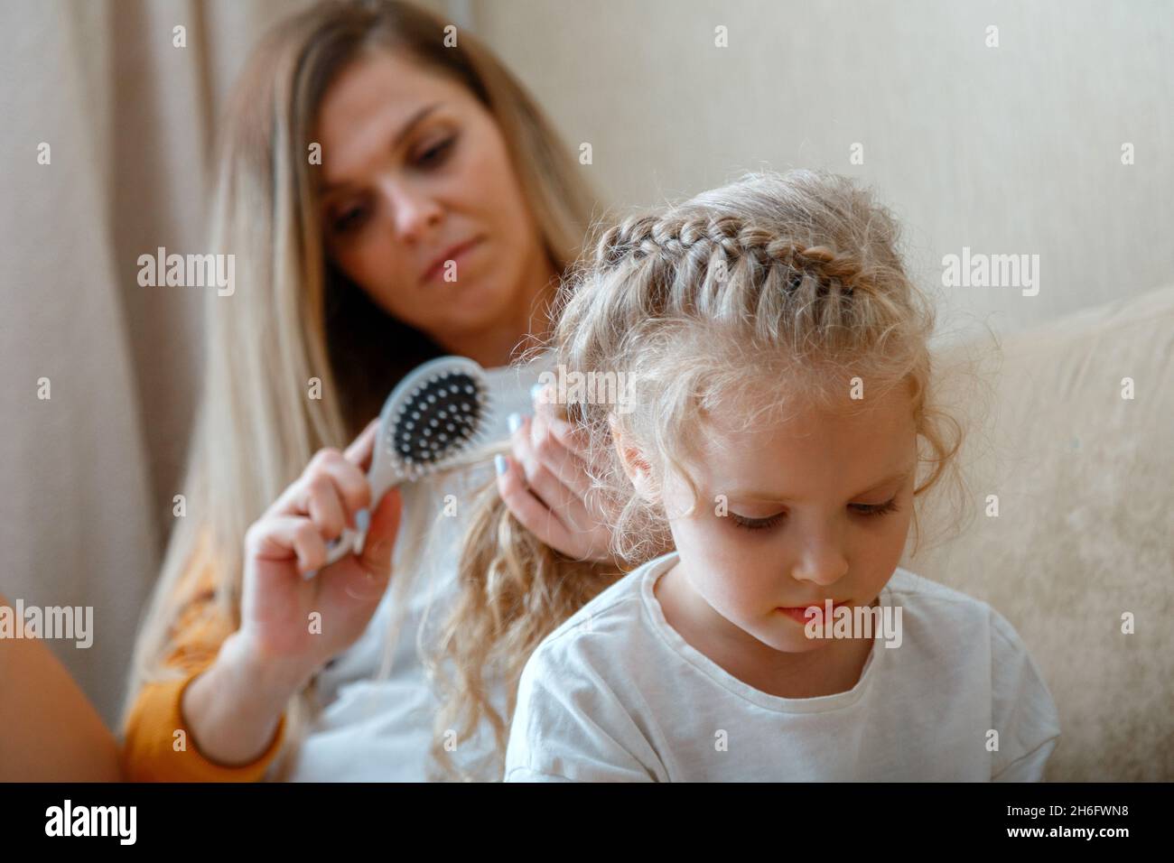 Mother combing daughter long hair by hairbrush. Caucasian blonde mom combing curly blond kid chaild. Mom does hair hairstyle and braids her braid Stock Photo