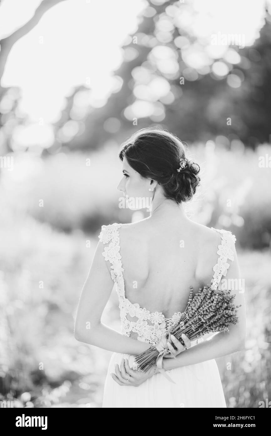 Sibenik, Croatia - 05.06.17: Bride in a lace white wedding dress stands with a bouquet of lavender behind her back, turning her head to the left Stock Photo