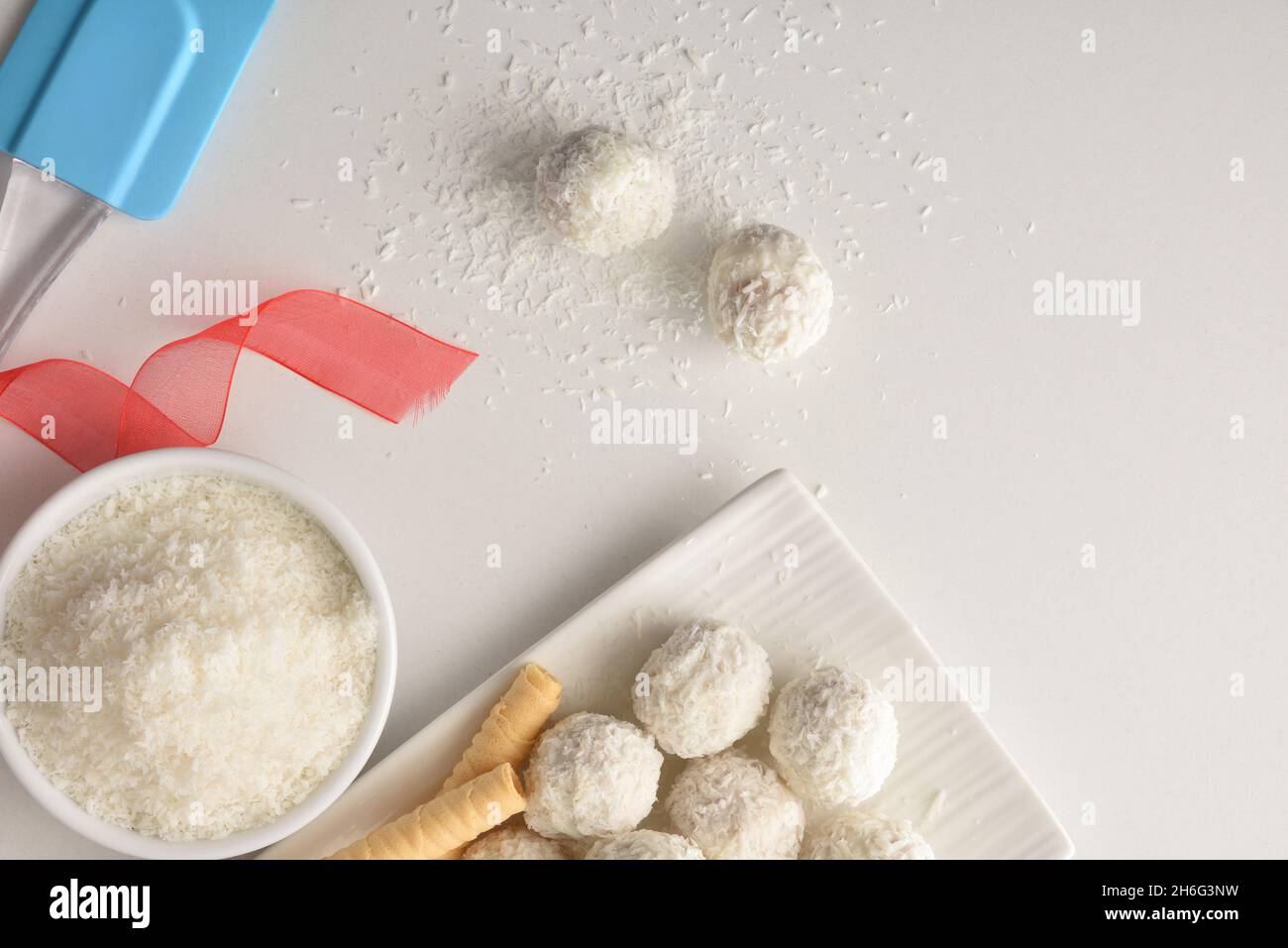 White kitchen table with coconut balls freshly made with bowl with grated coconut and kitchen tools and ribbon . Top view. Horizontal composition. Stock Photo