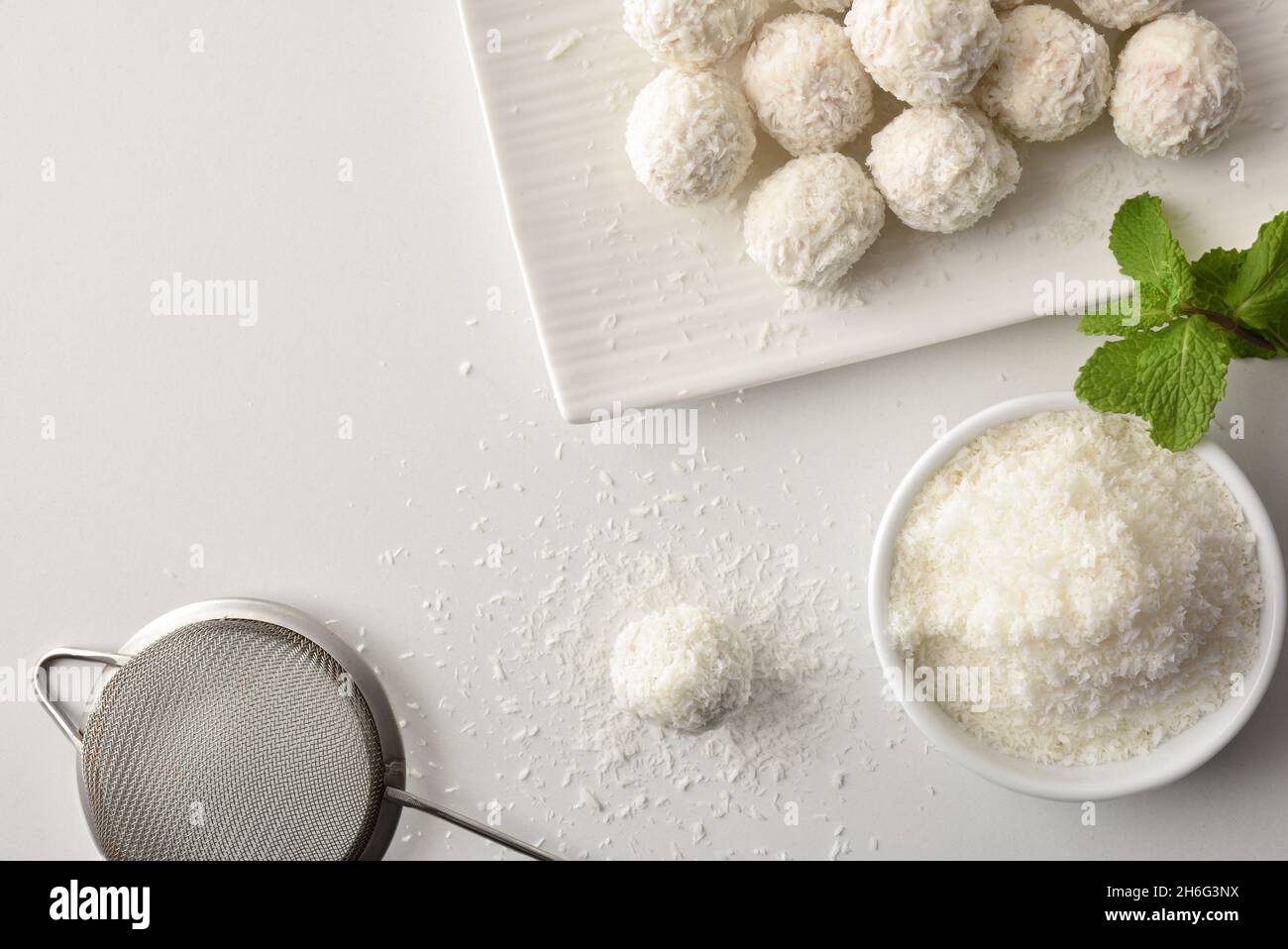 White kitchen table with coconut balls freshly made with bowl with grated coconut and kitchen tools. Top view. Horizontal composition. Stock Photo