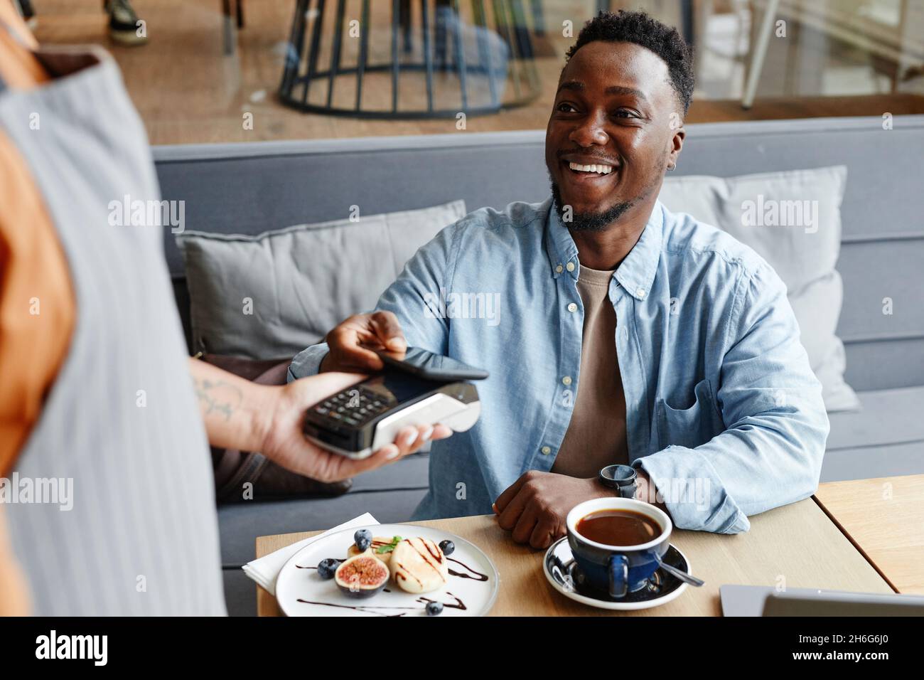 Joyful young Black man sitting at table in cozy cafe paying for lunch using his smartphone Stock Photo