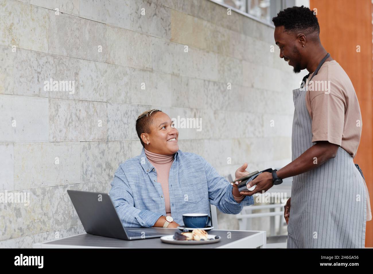 Joyful young Black woman sitting at table outdoor cafe paying for lunch using her smartphone Stock Photo