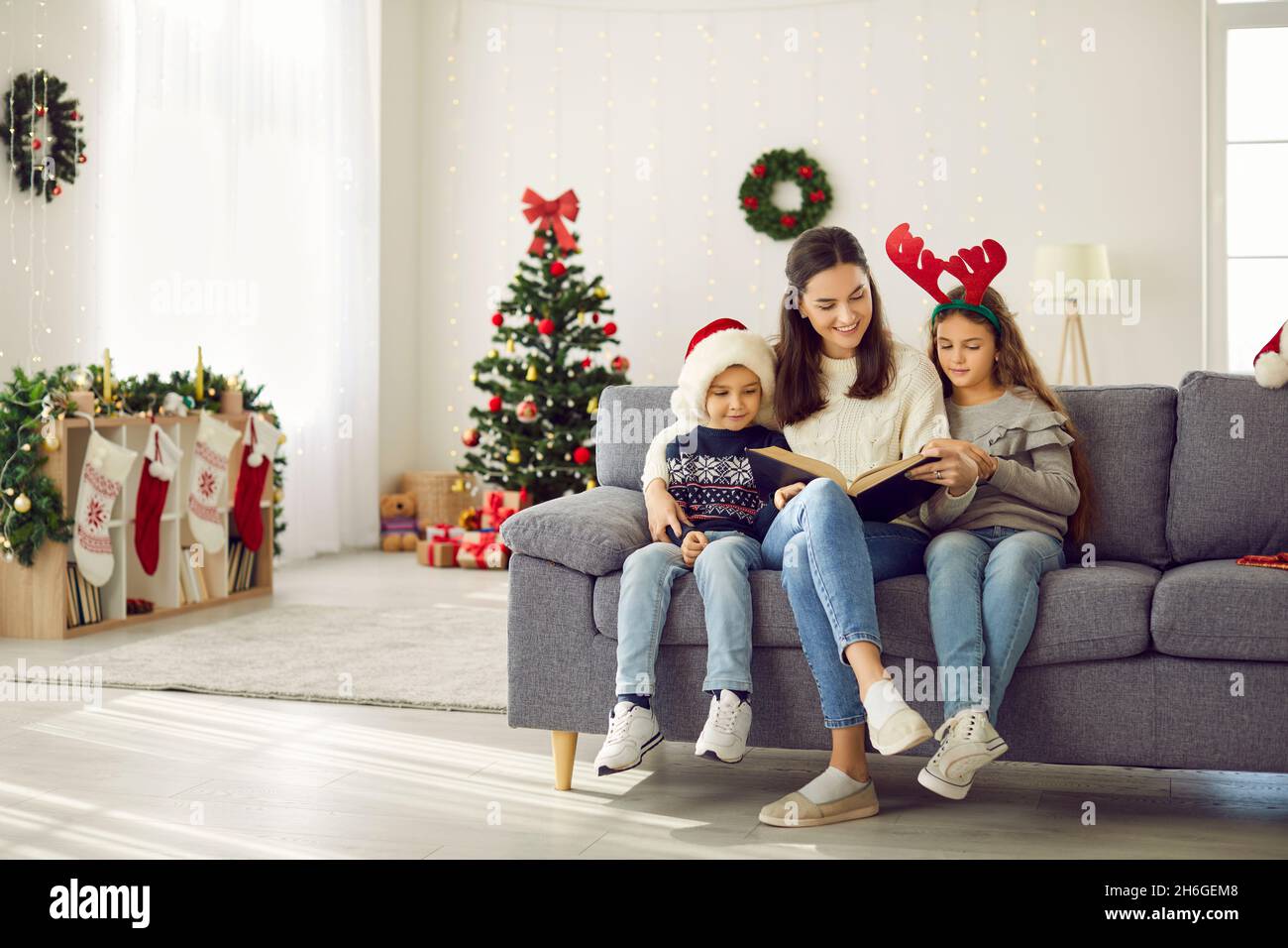Happy mom and kids reading book sitting on sofa in living room decorated for Christmas Stock Photo