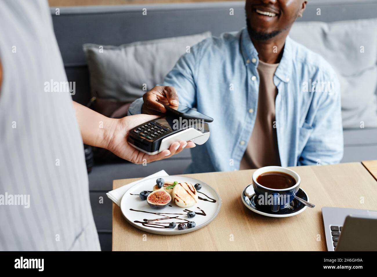 Unrecognizable young Black man sitting at table in modern cafe paying for order using his smartphone Stock Photo