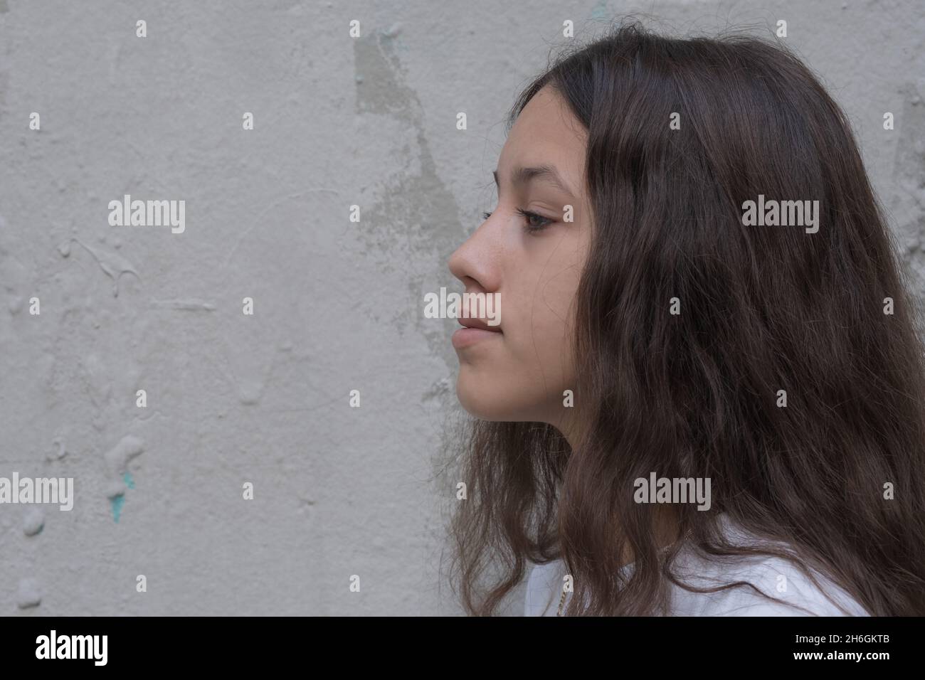 Close up profile of a teenage girl with long brown hair looks forward with a thoughtful expression. Background of gray wall, copy free space for text. Stock Photo