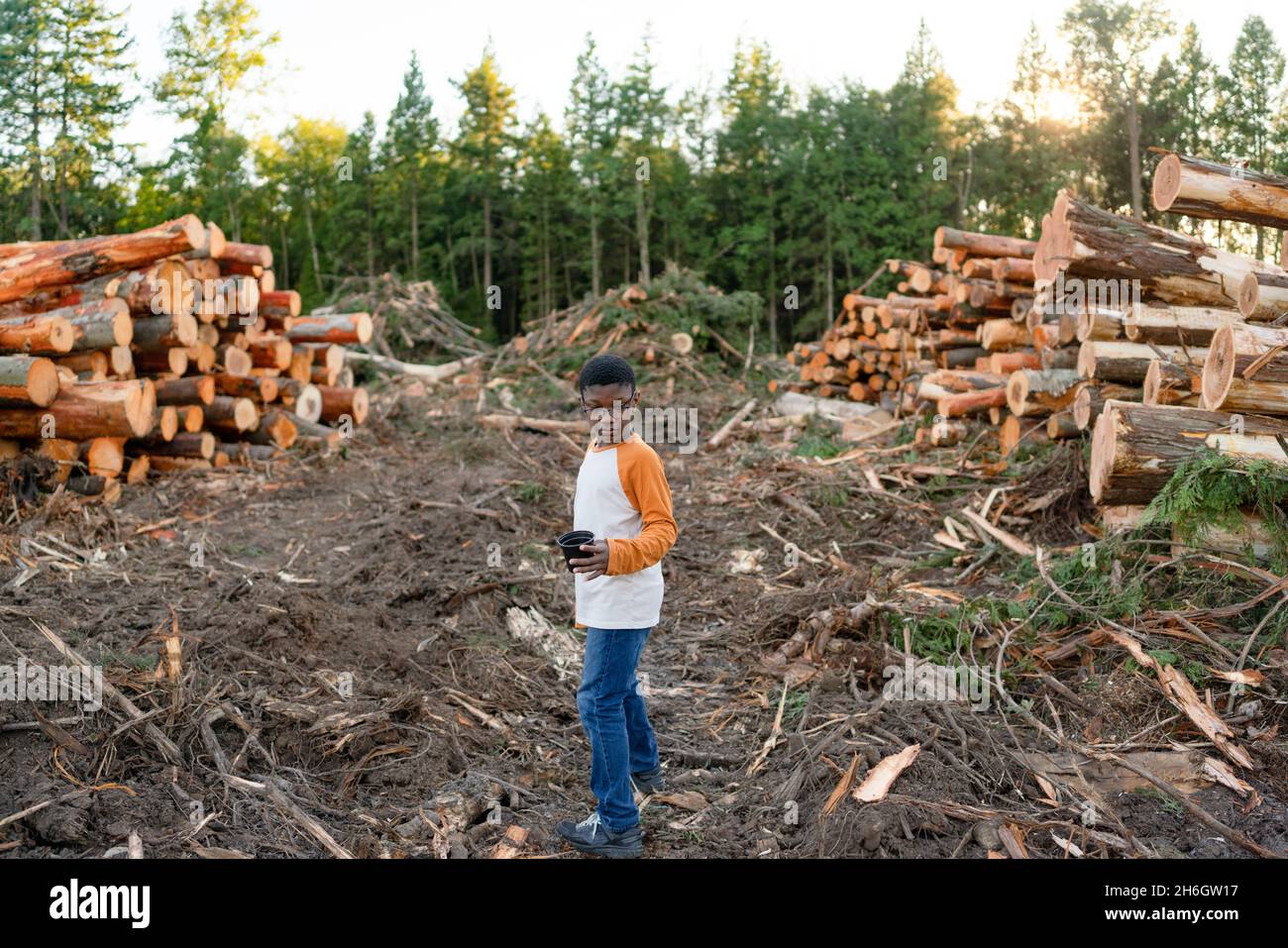 Young black boy stands alone on logging site surrounded by stacked logs and muddy ground. Stock Photo