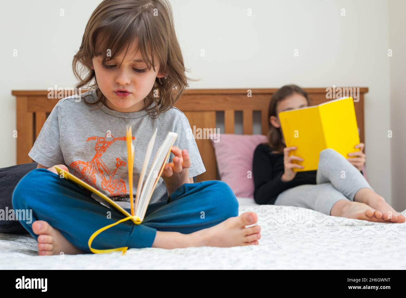 Young school age girl with her brother sitting on bed in bedroom and reading from yellow textbook. Stock Photo