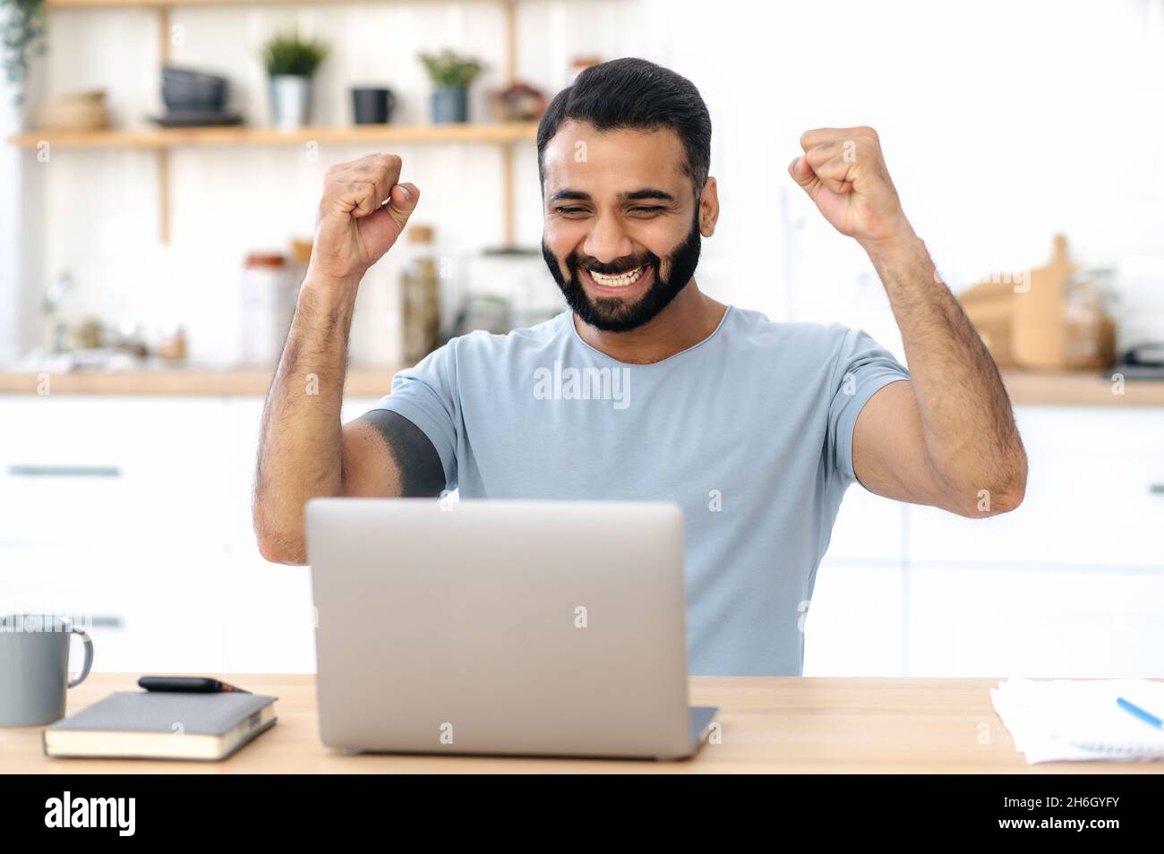 Happy excited lucky Indian man, in casual wear, freelancer, sits at a desk in kitchen, work from home, gesturing with his fists rejoicing in victory, success, good deal, big profit Stock Photo