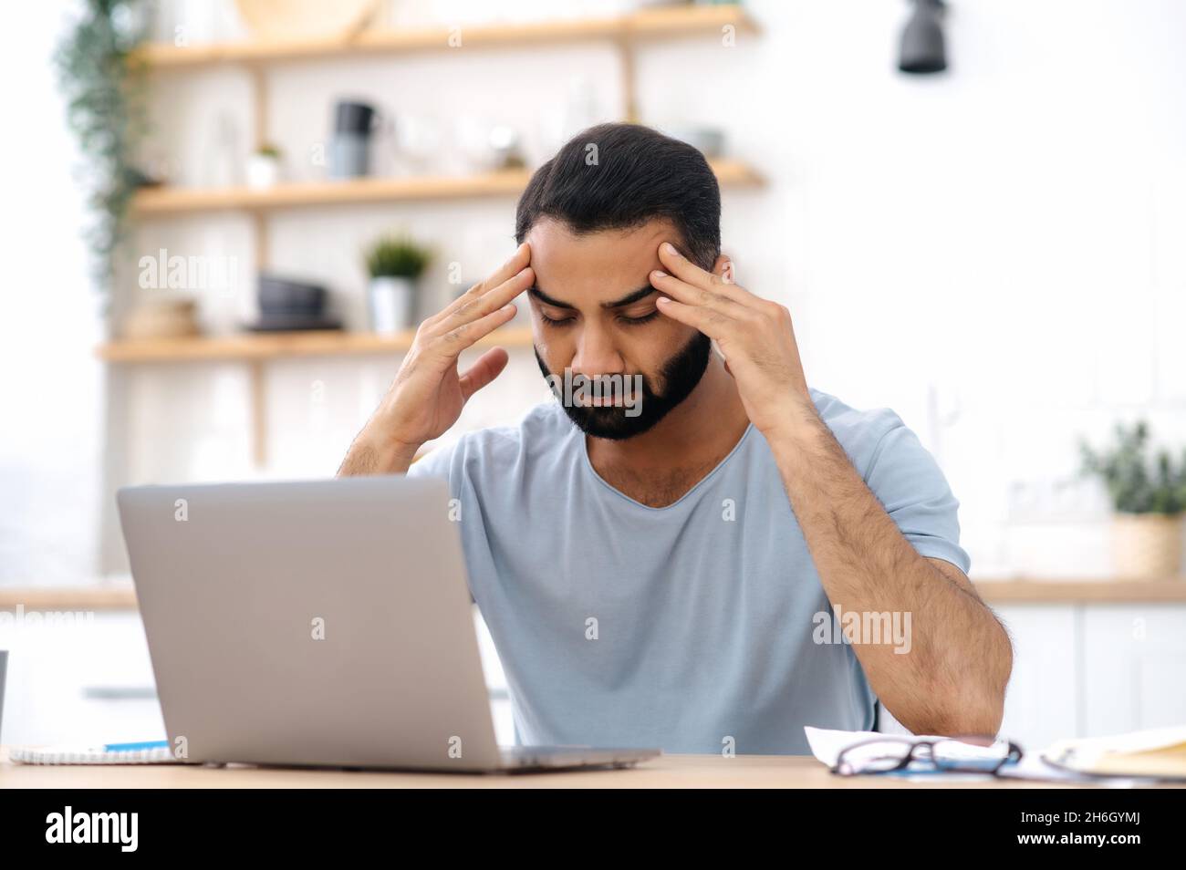 Headache, tiredness. A man of Indian nationality, a freelancer, works from home while sitting in the kitchen, uses a laptop, is tired, takes a break from work, closed his eyes, massages temples Stock Photo