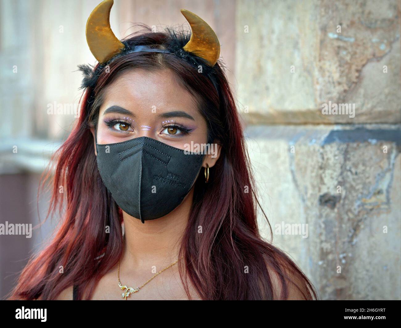 Pretty Mexican girl with yellow devil horns headband wears a black face mask during the global coronavirus pandemic and looks at the viewer. Stock Photo