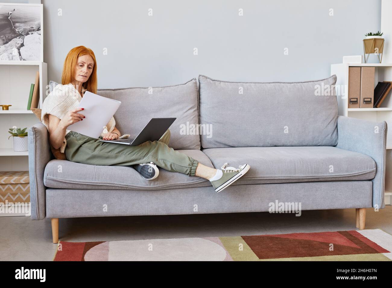 Full length portrait of adult red haired woman using laptop on sofa while working from home, copy space Stock Photo