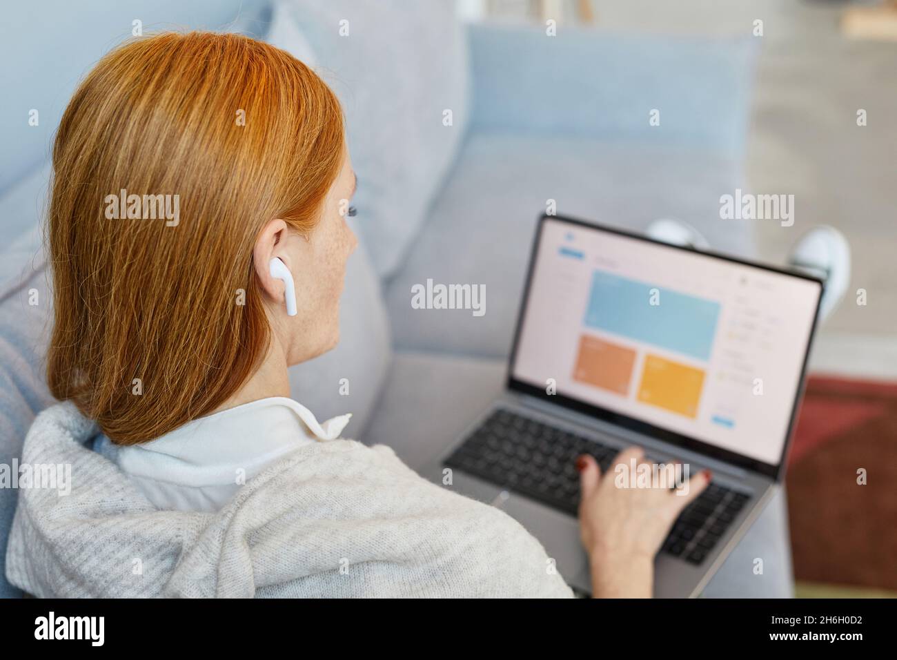 Back view portrait of adult red haired woman using laptop on sofa while studying online from home, copy space Stock Photo