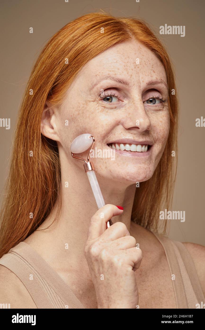 Vertical portrait of beautiful red haired woman with freckles smiling and doing face massage using roller Stock Photo