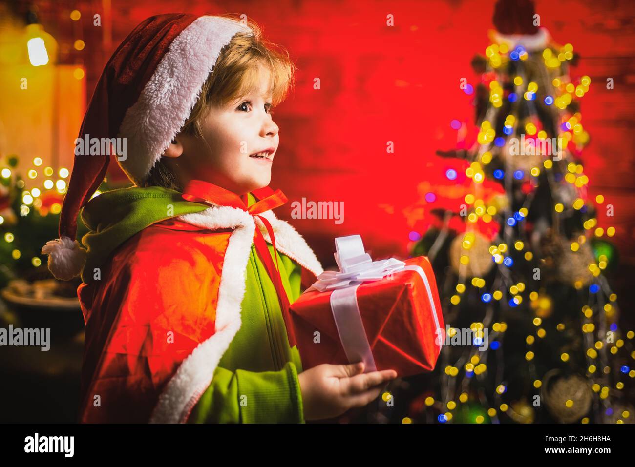 Happy little boy by near Christmas tree with Christmas gift. Little kid is wearing Santa clothes. Christmas kids concept. Stock Photo