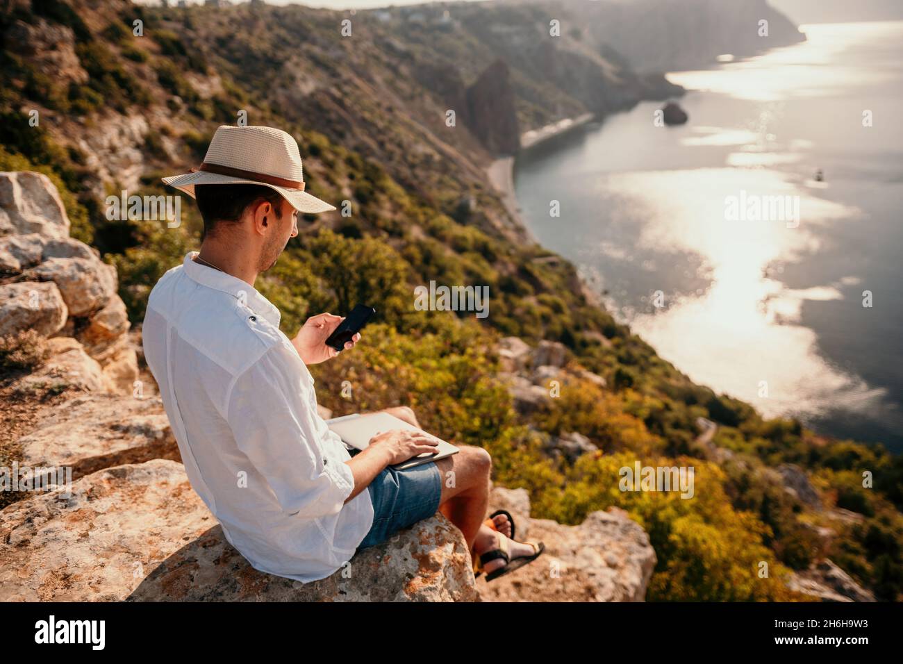 Digital nomad, man in the hat, a businessman with a laptop sits on the rocks by the sea during sunset, makes a business transaction online from a Stock Photo