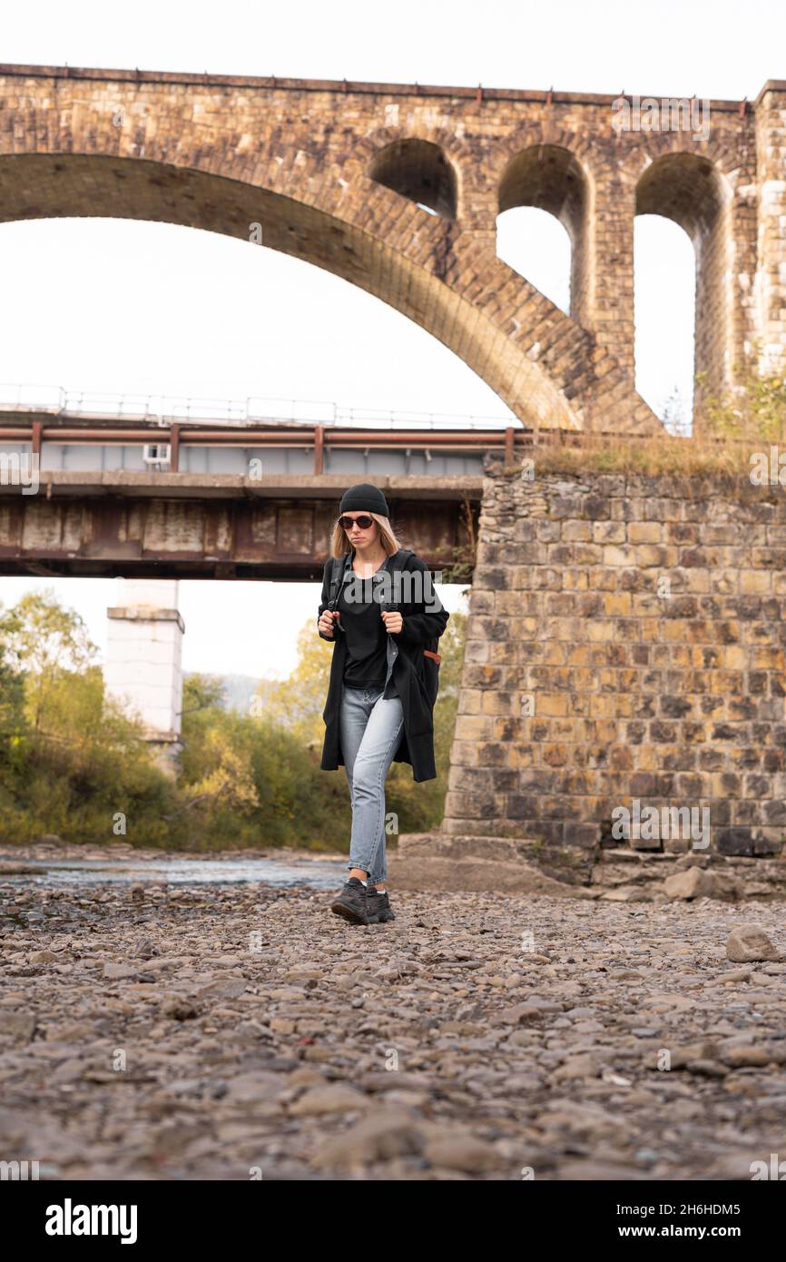 Arched old stone bridge, girl tourist walking by the river, hiking trekking, railway bridge background, tourism and travel. Stock Photo