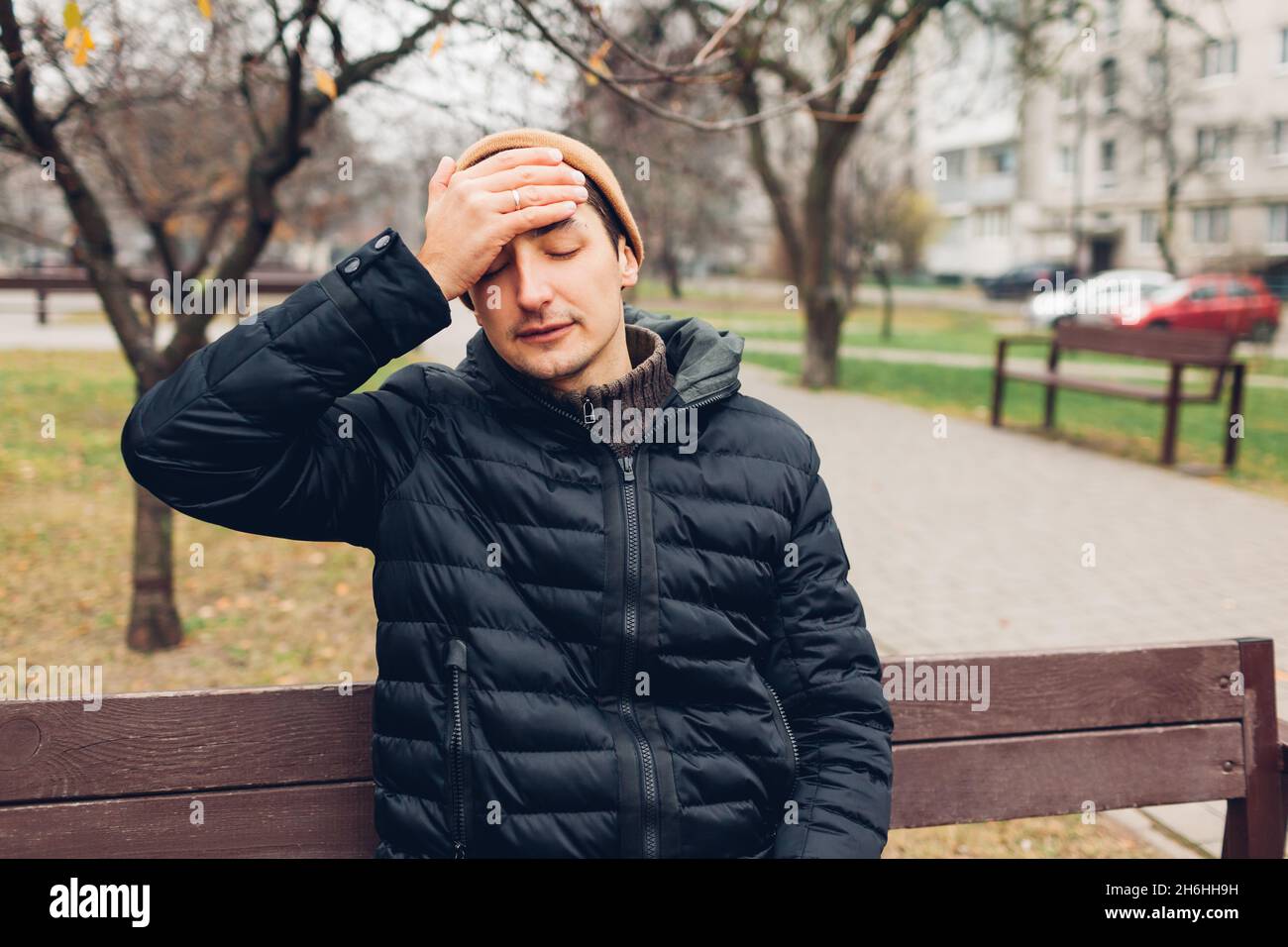 Sick man having headache and fever sitting on bench in fall park touching head. Coronavirus covid-19 pandemic flu. Helthcare during autumn Stock Photo