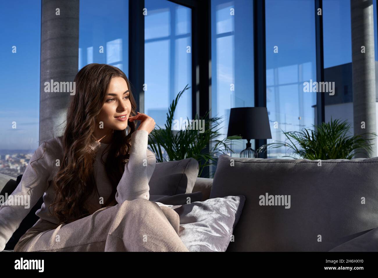 Pleasant young lady with long wavy hair smiling and looking aside while relaxing on cozy sofa with panoramic windows on background. Domestic lifestyle. Stock Photo