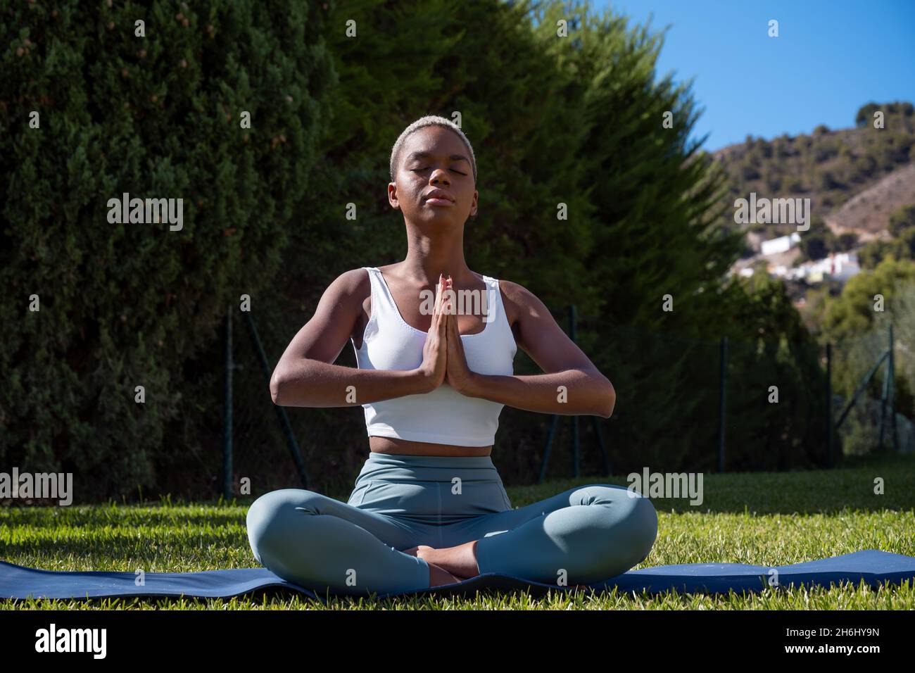 Tranquil African American female with short hair sitting in Lotus pose with prayer hands and closed eyes while doing yoga on mat on lawn in summer Stock Photo