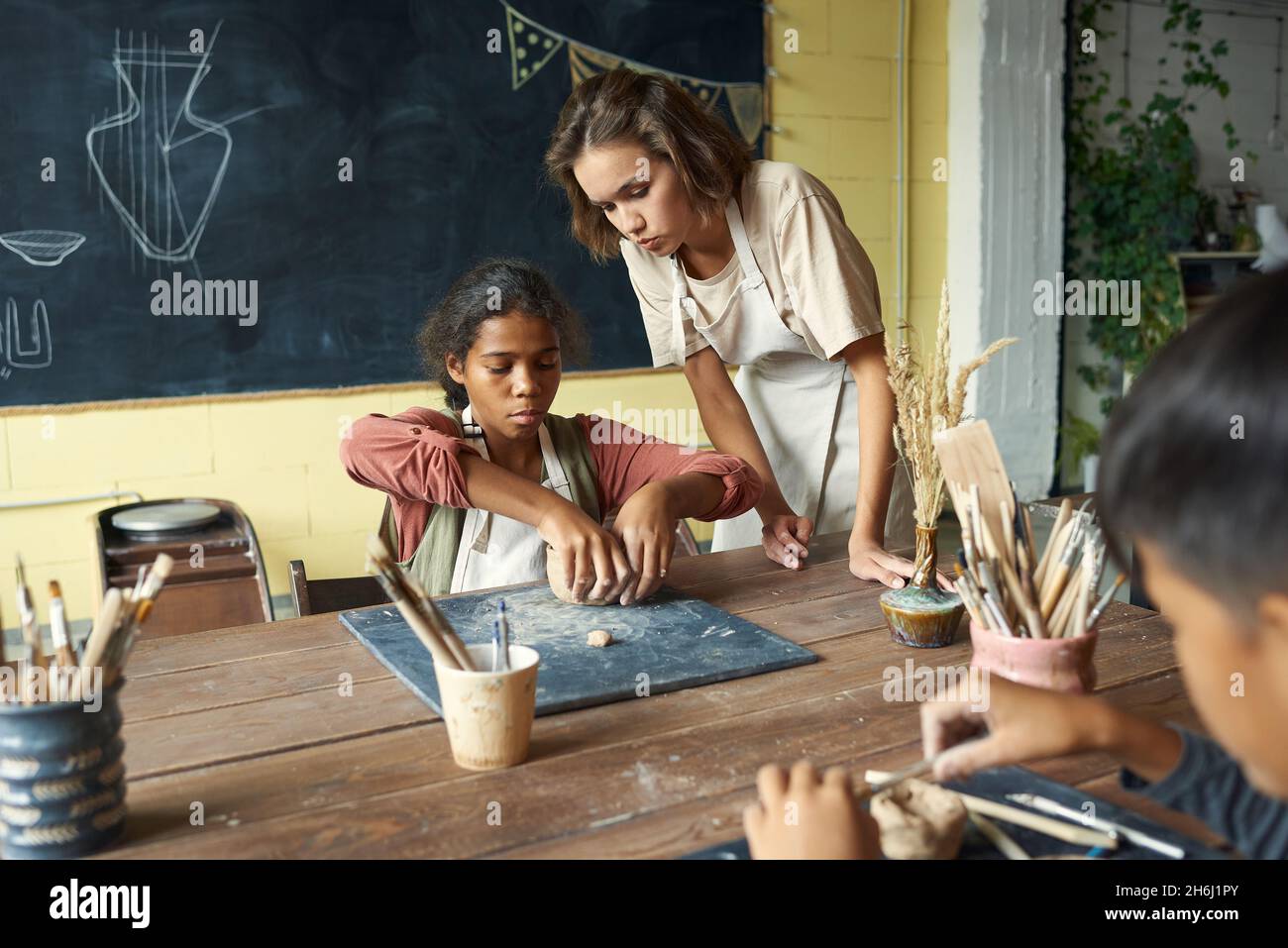 Young teacher of handcraft helping one of pupils at lesson while bending over African schoolgirl making earthenware Stock Photo