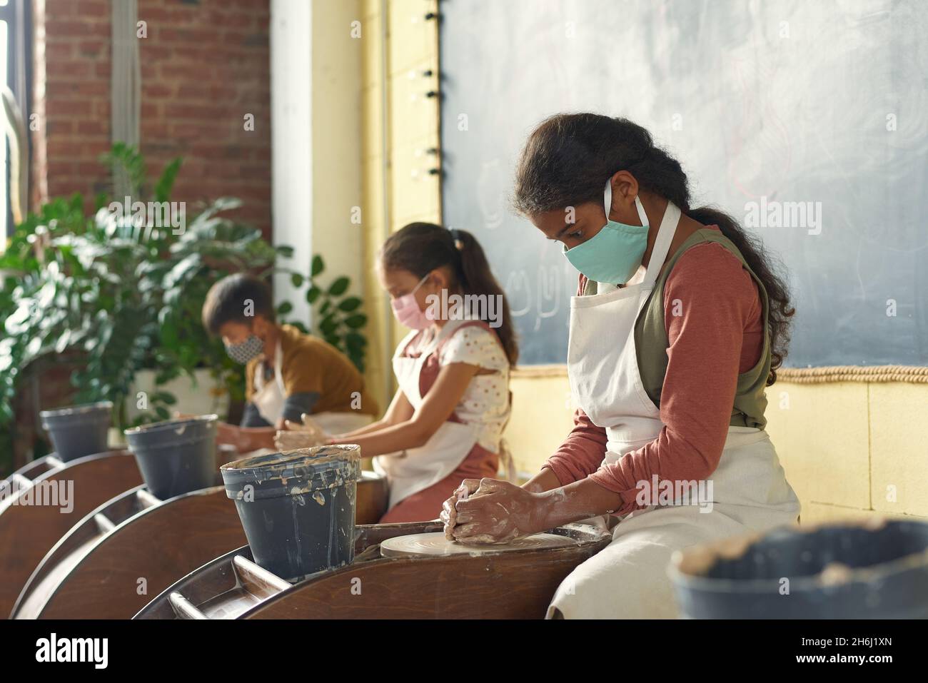 Two intercultural schoolgirls and schoolboy in aprons and protective masks sculpting clay jugs while attending pottery class Stock Photo