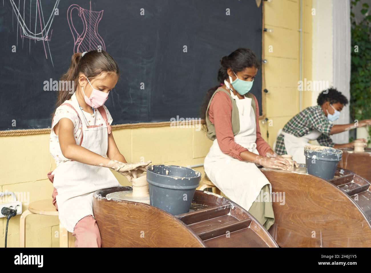 Row of pupils sitting along blackboard and sculpting clay pots in front of pottery wheels in large classroom or workshop Stock Photo