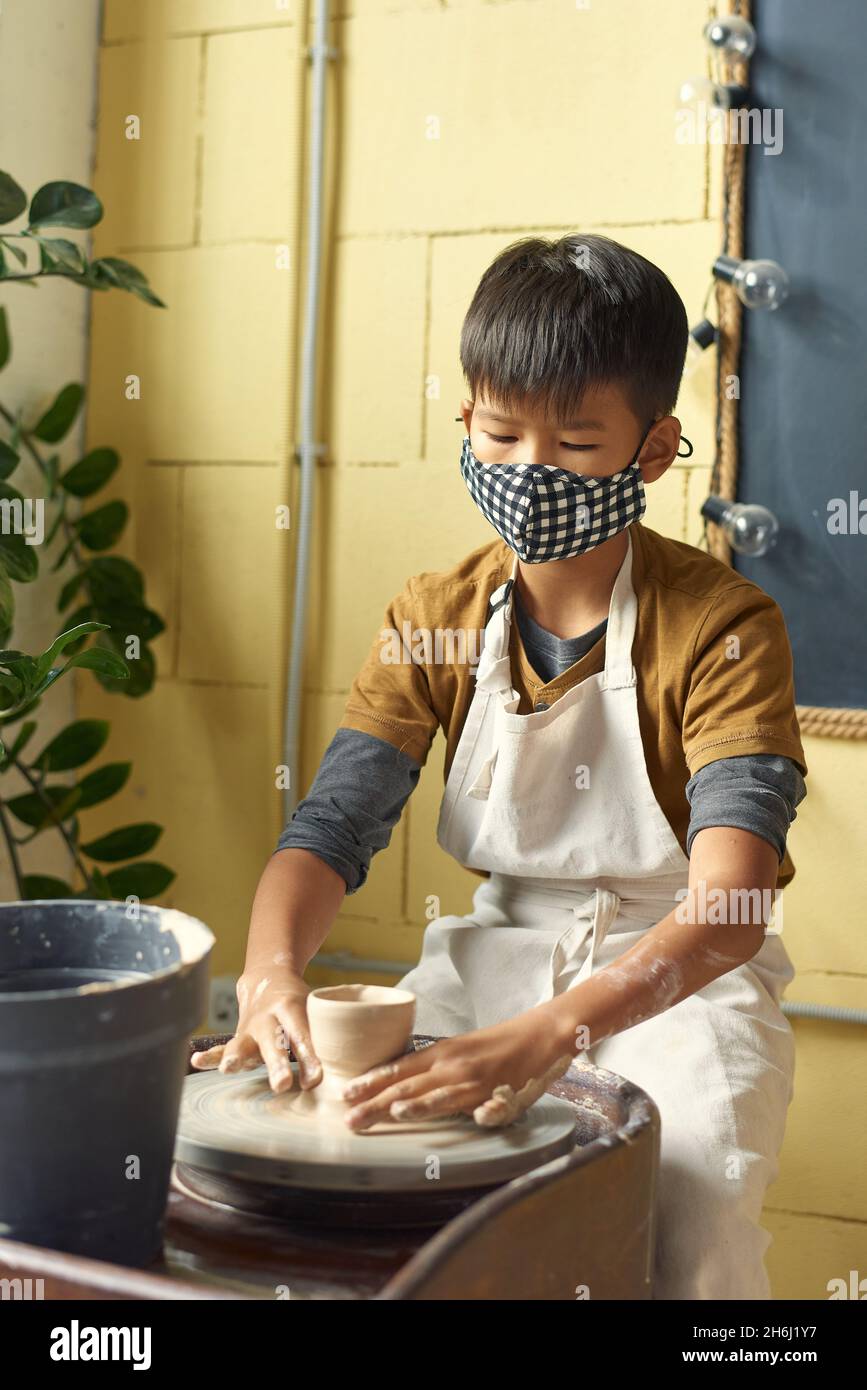 Diligent Asian schoolboy in protective mask sculpting clay pot at lesson of handcraft while sitting by pottery wheel Stock Photo