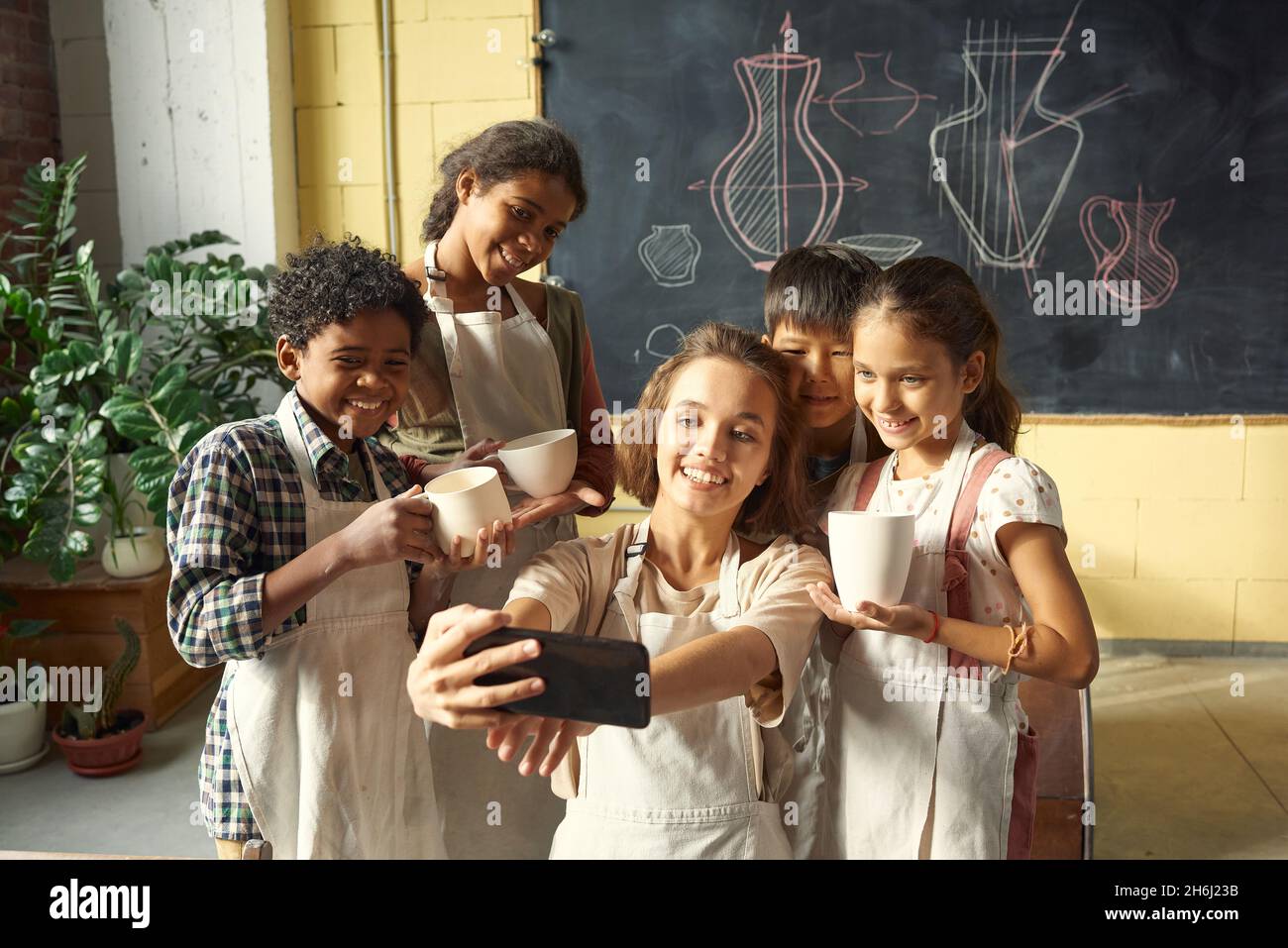 Happy young pottery craft teacher making selfie with group of diligent learners holding handmade clay mugs Stock Photo