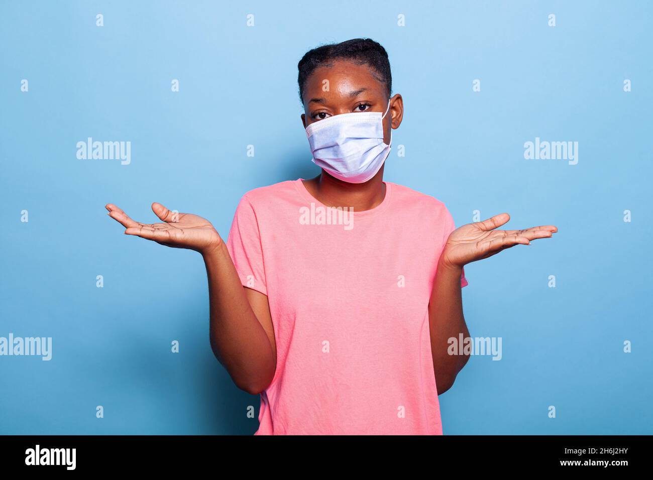 Confuse african american teenager with protective face mask against coronavirus shrugging shoulders looking at camera posing over blue background in studio. Uncertain question gesture Stock Photo