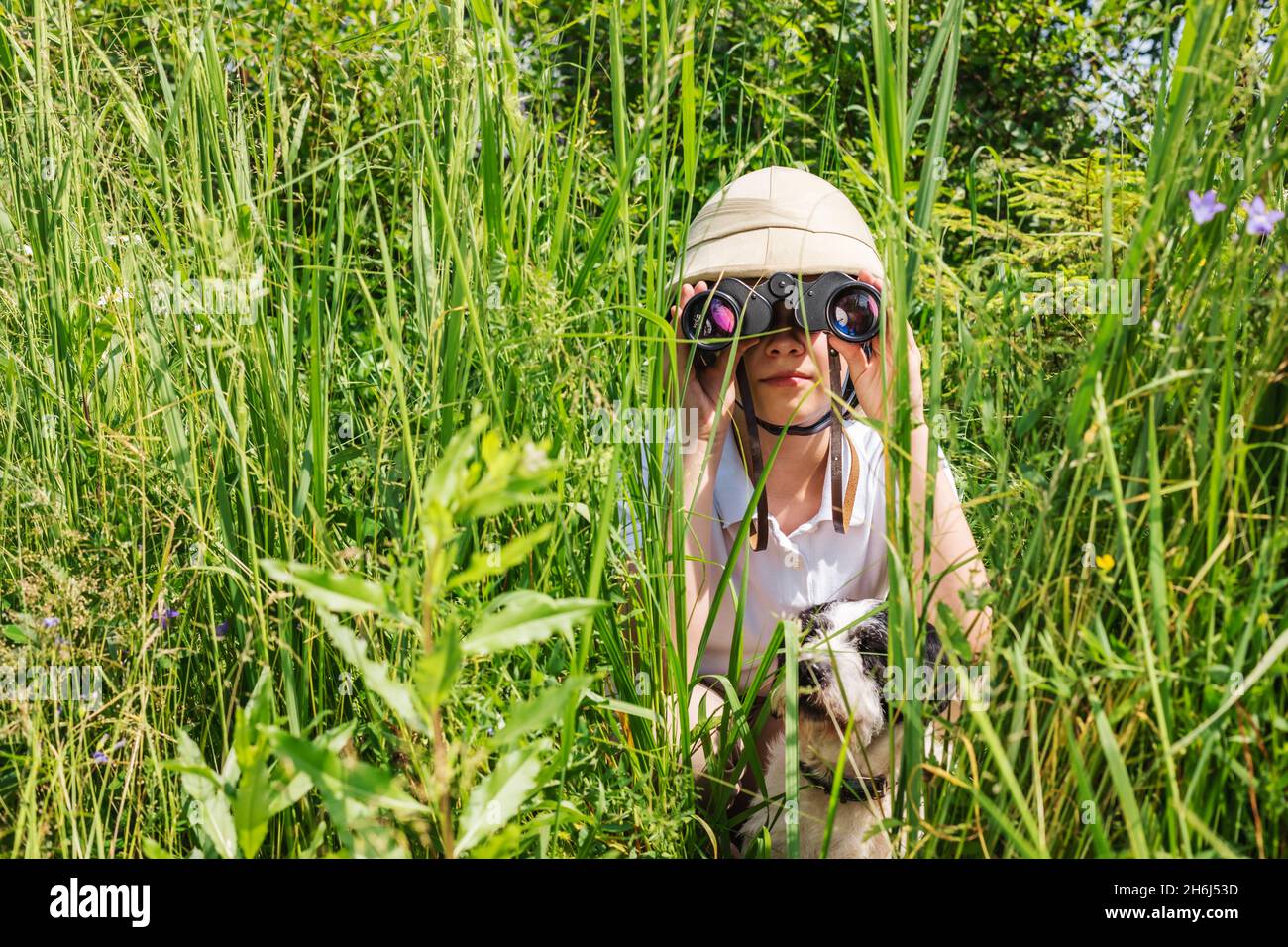Preteen girl wearing cork helmet hiding in the grass with her dog looking through binoculars Stock Photo