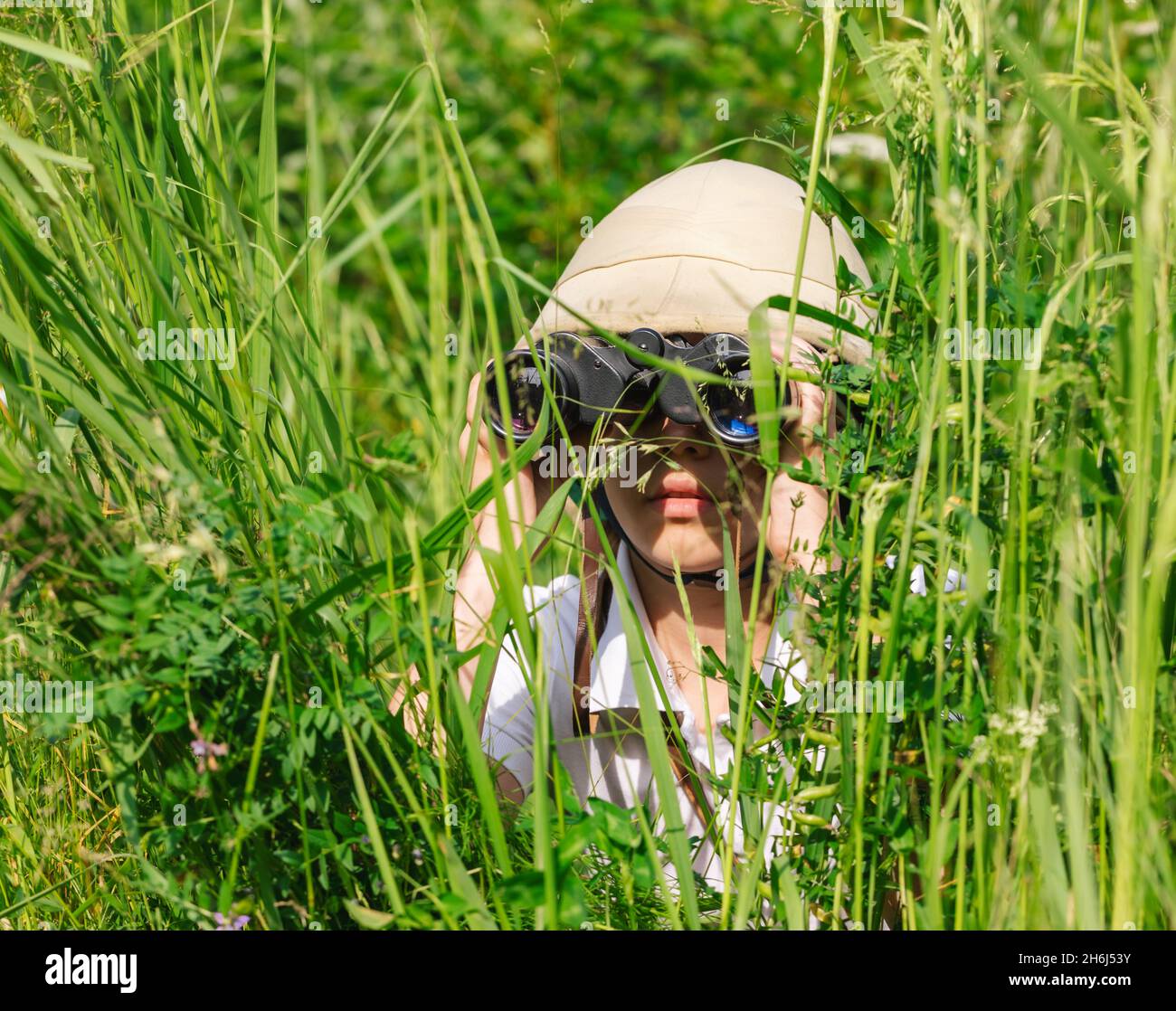 Preteen girl wearing cork helmet hiding in the grass looking through binoculars. Discovery concept Stock Photo