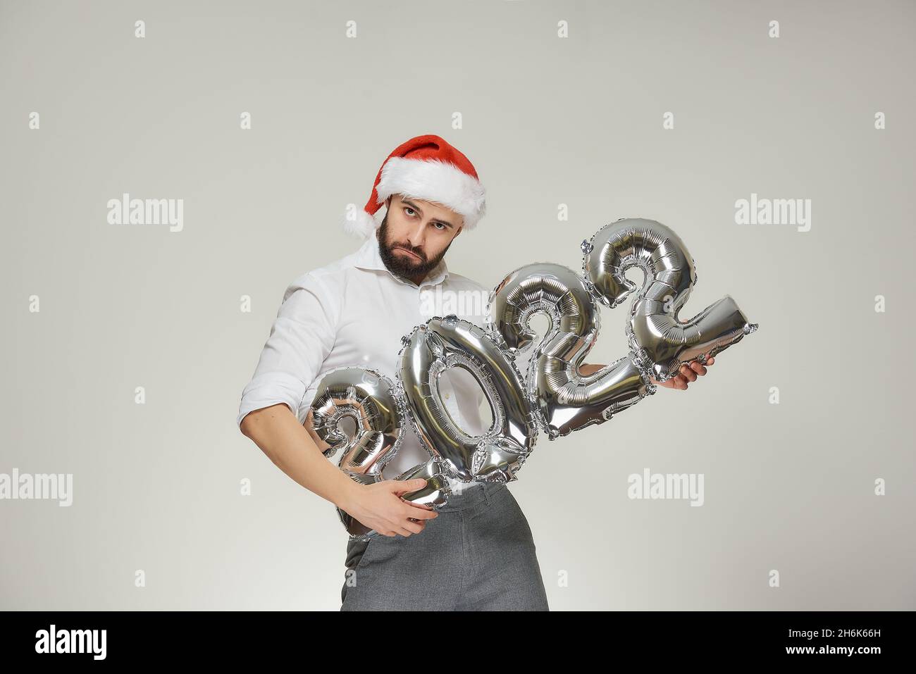 A tough man in a red velvet Santa hat is holding silver balloons in the shape of 2022 like a machine gun. A guy with a beard at a New year party. Stock Photo