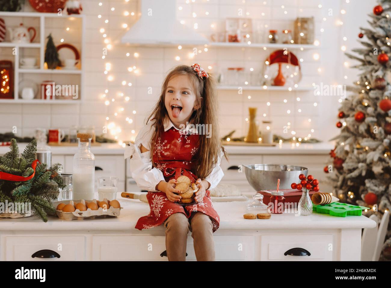 A happy little girl in the Christmas kitchen is sitting on the table with cookies in her hands and making faces. Stock Photo