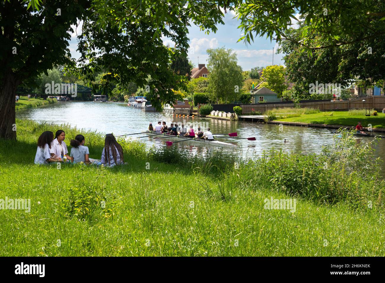 Girls sitting together under tree on grass beside the River Cam near Cambridge with rowers rowing on Downing college eight on river Cambridge England Stock Photo