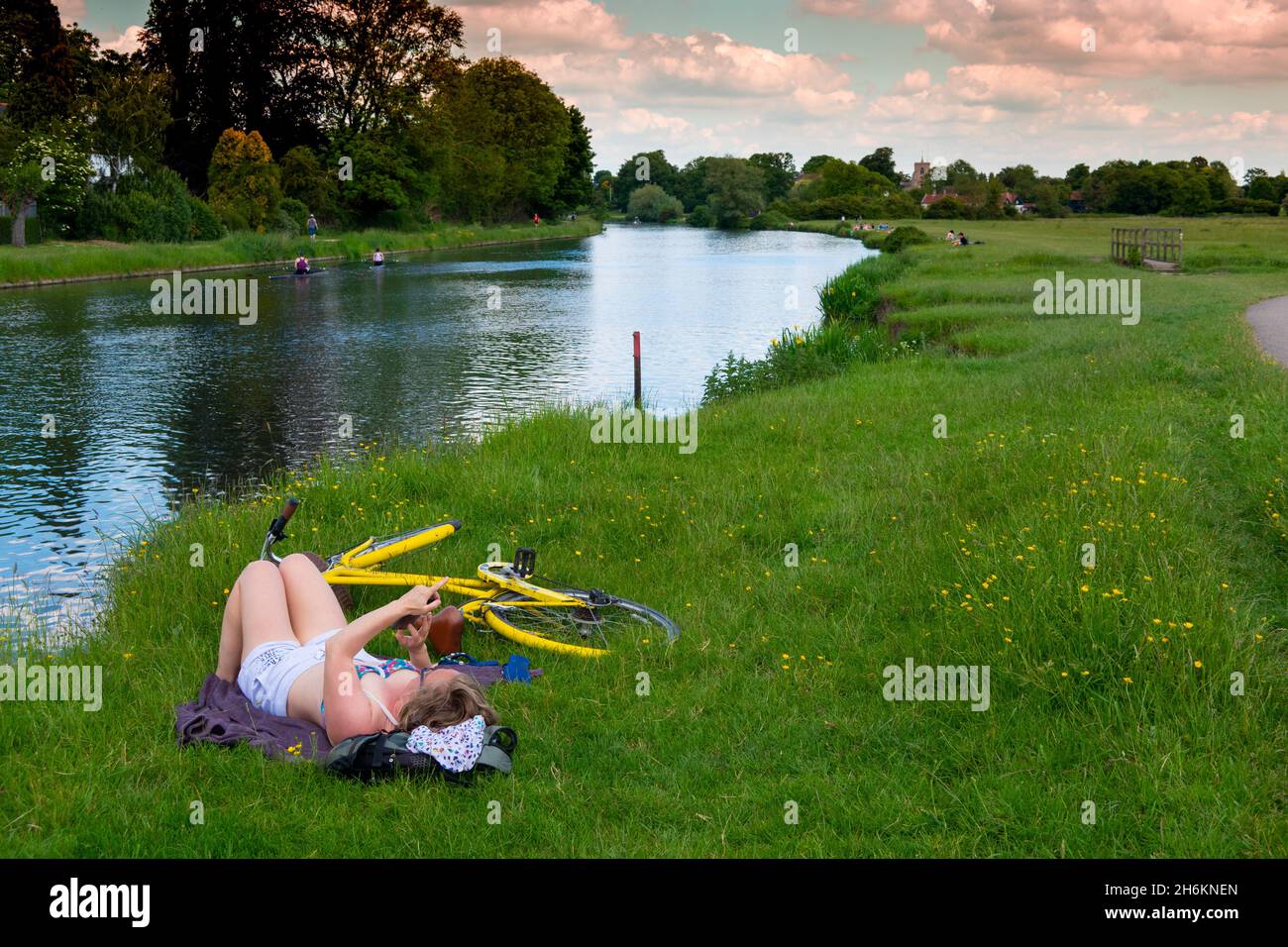 Young girl relaxing alone lying on grass meadows looking at her phone with yellow cycle on the bank of the River Cam near Cambridge England Stock Photo