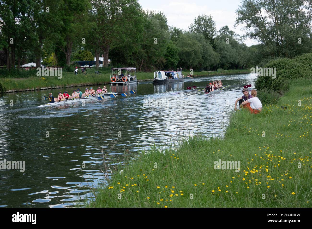 Couple sitting relaxing on river bank beside the River Cam near Cambridge with rowers on eight on river Stock Photo
