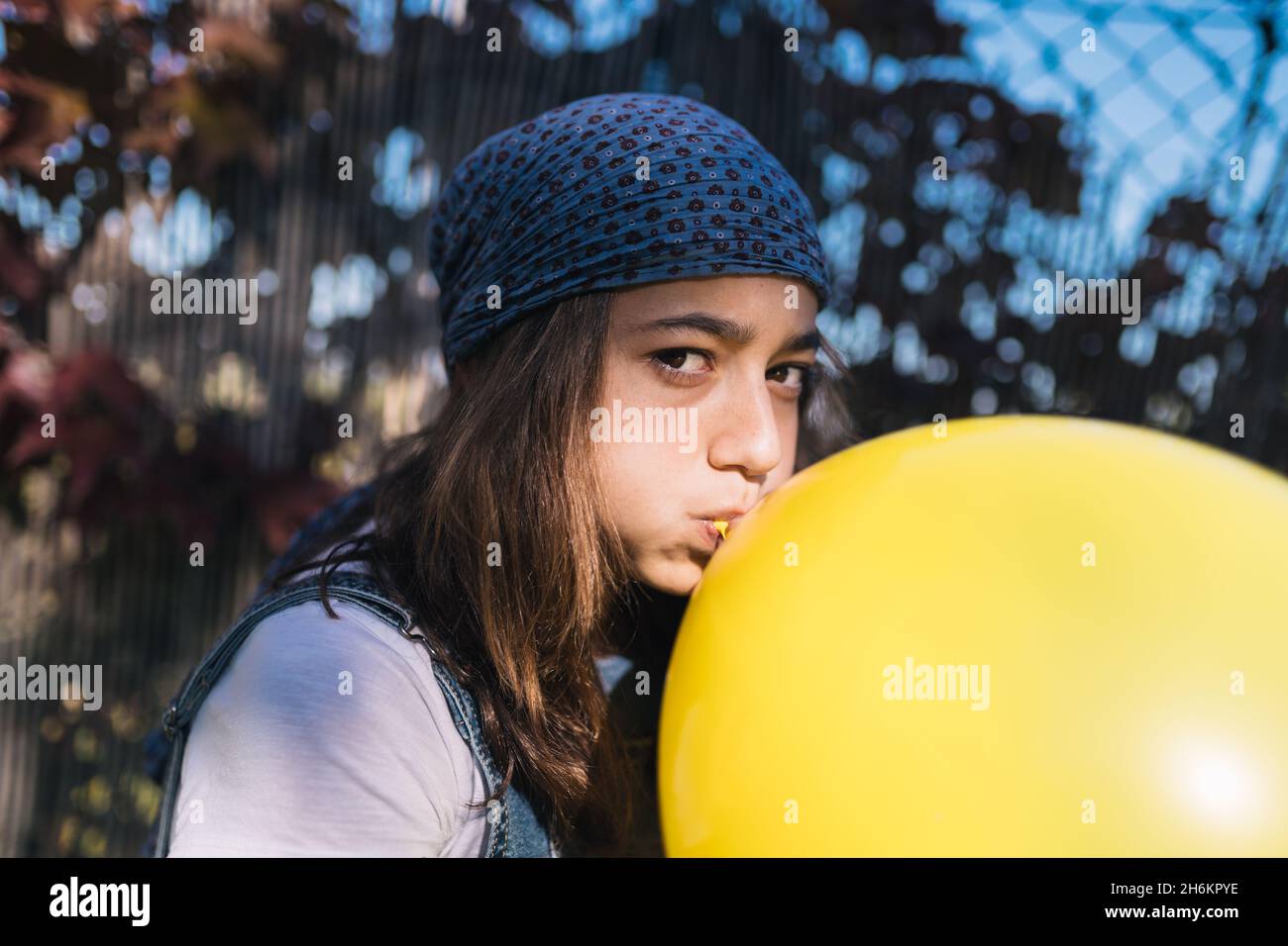 Teenage girl with a headscarf and long brown hair blowing up a huge yellow balloon sitting in a green garden on a sunny day. Adolescence Stock Photo