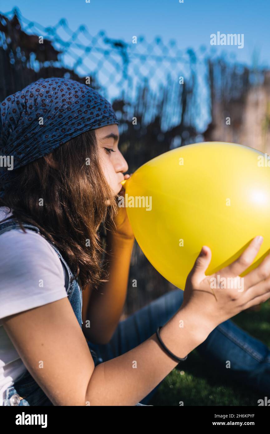 Teenage girl with a headscarf and long brown hair blowing up a huge yellow balloon sitting in a green garden on a sunny day. Adolescence Stock Photo