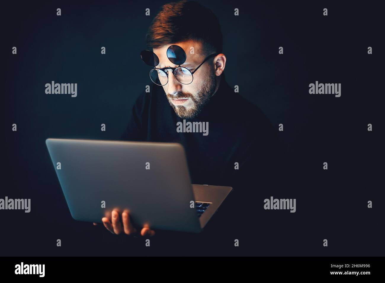 Young bearded hipster man wearing glasses while using laptop over black studio background  Stock Photo