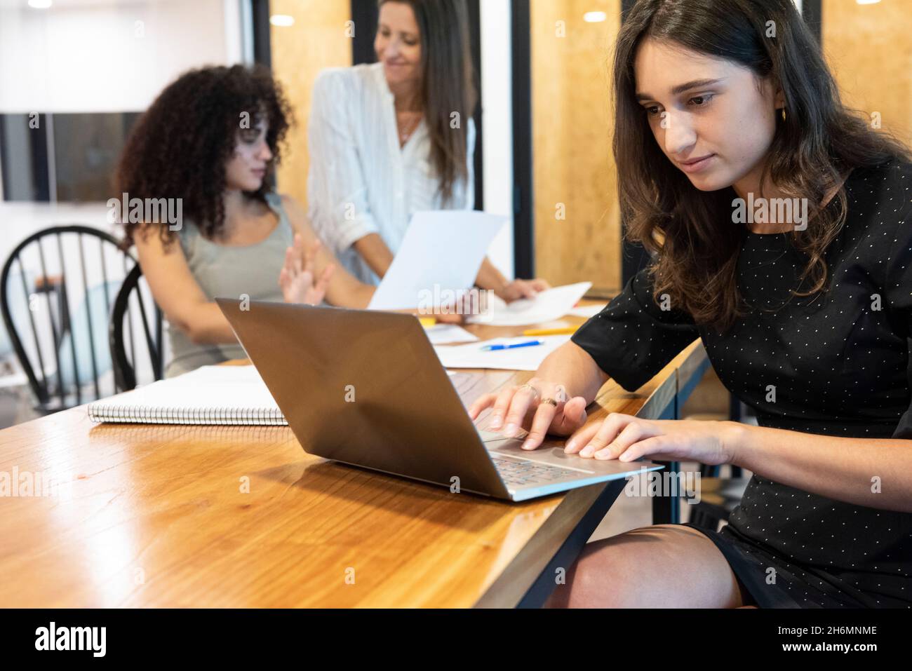 Young woman working on laptop with businesswomen discussing in background Stock Photo