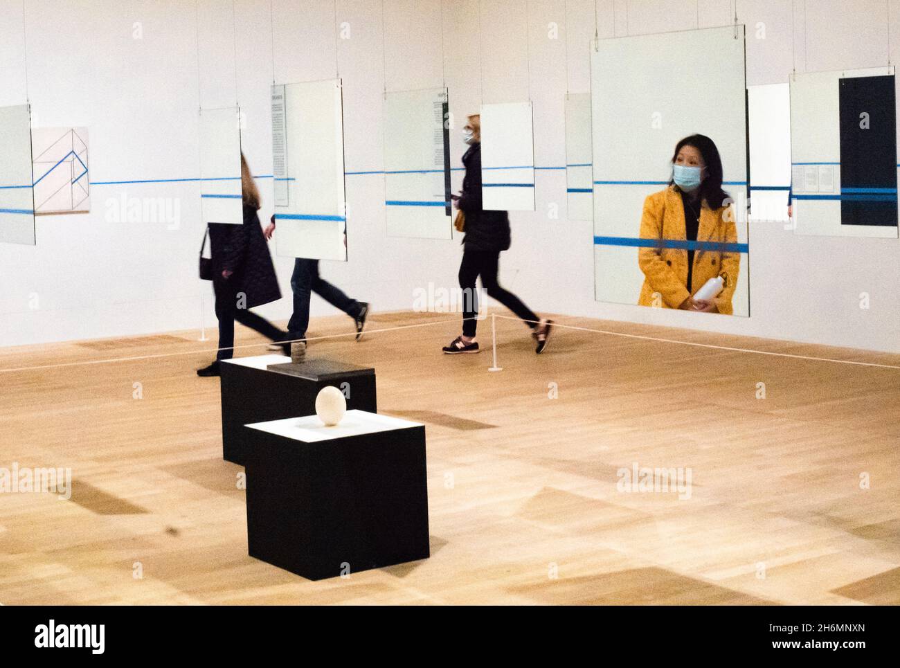 An Asian woman wearing a surgical mask is seen in a mirror as others walk past in a gallery showing an art installation at Tate Modern Stock Photo